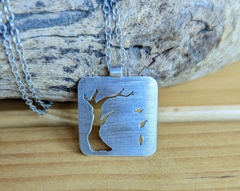 May include: A silver necklace with a square pendant featuring a cutout design of a tree with leaves falling. The pendant is hanging on a silver chain.