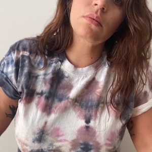 May include: A white tie-dye t-shirt with shades of pink, blue, and gray. The shirt has a relaxed fit and a crew neck.