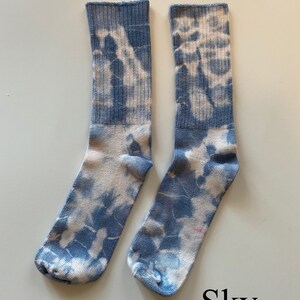May include: A pair of blue and white tie-dye socks. The socks have a light blue background with white tie-dye patterns. The socks are folded and laid out on a white surface. The word "Sky" is written in black below the socks.