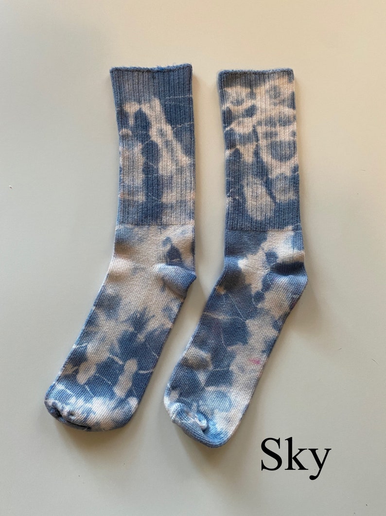 May include: A pair of blue and white tie-dye socks. The socks have a light blue background with white tie-dye patterns. The socks are folded and laid out on a white surface. The word "Sky" is written in black below the socks.