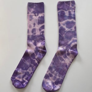May include: A pair of purple and white tie-dye socks. The socks have a light purple base with white tie-dye swirls. The socks are labeled "Violet".