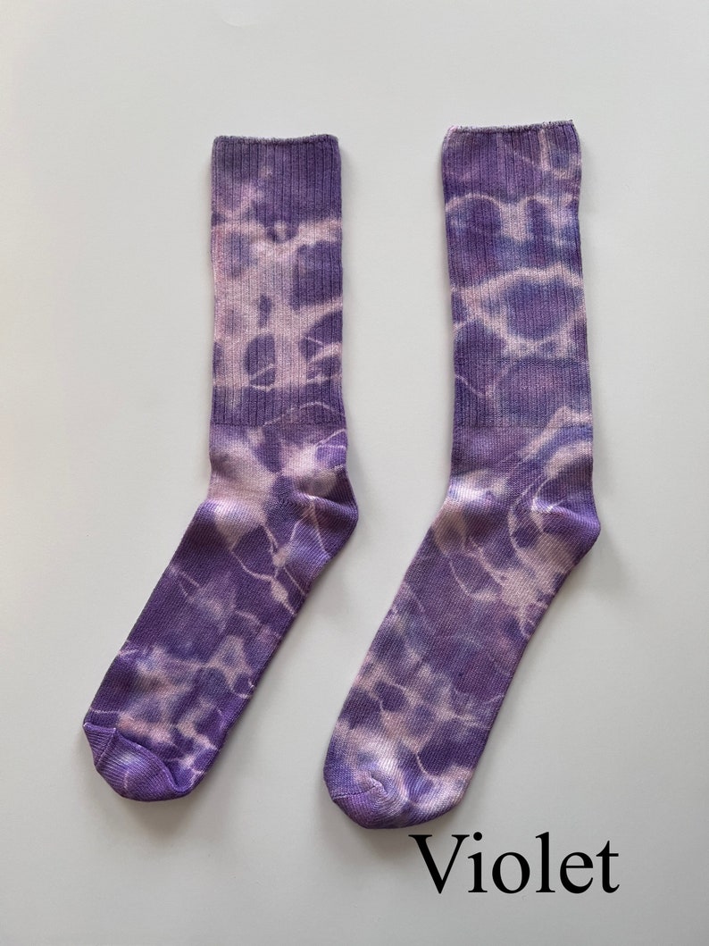 May include: A pair of purple and white tie-dye socks. The socks have a light purple base with white tie-dye swirls. The socks are labeled "Violet".