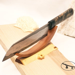 May include: A wooden knife stand with a curved wooden base and a black and brown handle knife resting on it. The knife has a textured blade and a wooden cutting board is in the background.