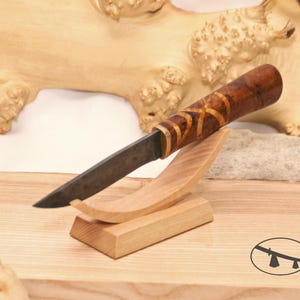 May include: A small, handcrafted knife with a dark brown wooden handle and a black blade. The knife is displayed on a wooden stand.