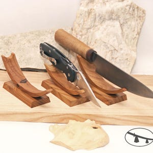 May include: Three wooden knife stands, each holding a different knife. A large, curved knife with a wooden handle rests on one stand. A folding knife and a small wooden carving are also present.