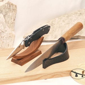 May include: A folding knife with a black handle and a silver blade resting on a curved wooden stand, alongside a fixed-blade knife with a light brown wooden handle and a silver blade resting on a black textured curved stand.