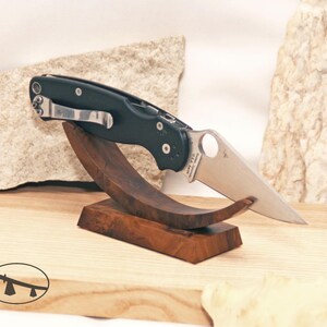 May include: A black folding knife with a silver blade rests on a custom-made wooden stand. The knife features a pocket clip and a partially serrated edge. The stand is made of dark wood and has a curved design, showcasing the knife.