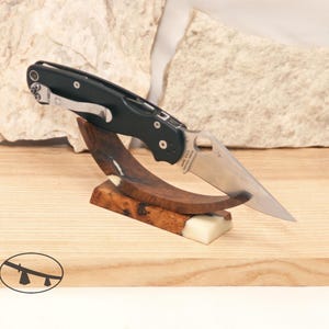 May include: A black and silver folding knife with a partially serrated blade rests on a custom wooden display stand. The stand is made of dark wood and a white resin base, showcasing the knife's design. The knife is a practical tool.