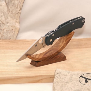 May include: A folding pocket knife with a silver-toned metal blade featuring a circular cutout and a black handle, resting on a curved, polished wooden display stand with a visible grain pattern. The blade displays a small spider logo engraving.