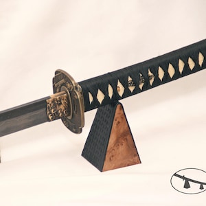 May include: A katana sword with a black handle and a decorative bronze guard. The blade has a dark, textured appearance. The handle is wrapped with black cord and features diamond-shaped accents. The sword rests on a wooden stand.