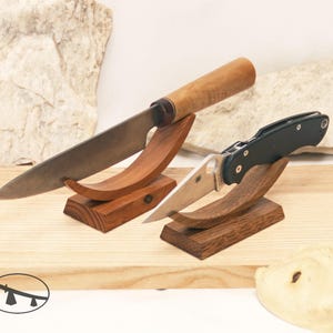 May include: Two wooden knife stands with curved cradles, one holding a fixed-blade knife with a light wood handle and the other holding a black folding knife with a silver blade. A small circular logo is visible in the bottom left corner.