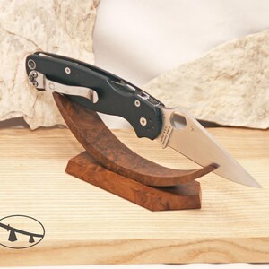 May include: A black folding knife with a silver blade is displayed on a wooden stand. The knife has a pocket clip and is set against a light-colored background. The stand is made of dark wood.