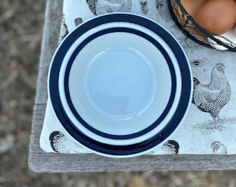 Könnte beinhalten: Eine blaue und weiße Keramikschüssel mit blauem Rand steht auf einer weißen Stoffoberfläche mit einem Hahnmuster.