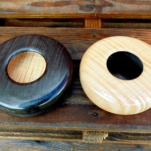 Puede incluir: Dos cuencos de madera con una abertura en forma de anillo en el centro. Un cuenco es negro con un anillo de madera clara, y el otro es de madera clara con un anillo negro.