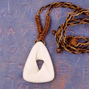 Op de afbeelding: Een crèmekleurige hangerketting met een driehoekig ontwerp en een centrale opening. De hanger hangt aan een gevlochten koord in bruine en bruin/beige tinten. De ketting wordt gepresenteerd op een blauw oppervlak.