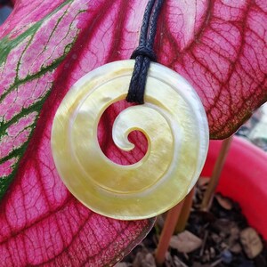 May include: A yellow and white shell pendant with a spiral design hangs from a black cord. The pendant is on a pink and green leaf.