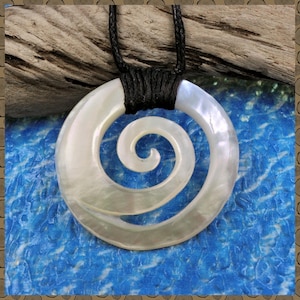 Pode incluir: Um colar com pingente com um pingente grande, circular, de madrepérola com um design em espiral. O pingente é suspenso por um cordão preto. O fundo é uma superfície azul texturizada com madeira flutuante.