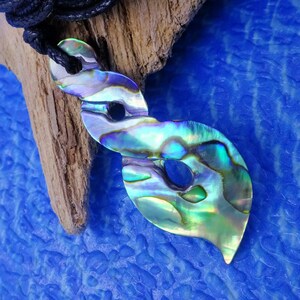 Può includere: Collana con pendente a forma di conchiglia di abalone iridescente con sfumature di blu, verde e viola. Il pendente è collegato a un cordino nero. La collana è esposta su un pezzo di legno alla deriva su sfondo blu.