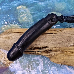 Può includere: Un ciondolo nero a forma di fallo in legno scolpito, sospeso a un cordino nero. Il ciondolo è liscio e arrotondato, con una punta dettagliata. Lo sfondo presenta un pezzo di legno trasportato dall'acqua su uno sfondo di acqua blu e bianca.