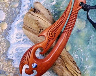 May include: Two intricately carved wooden pendants with a reddish-brown hue. One features a spiral design and a small abalone shell inlay. The other has a carved spiral pattern. Both are displayed on driftwood against a backdrop of a beach scene.