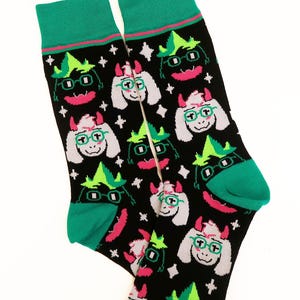 May include: A pair of black crew socks featuring a repeating pattern of a white, horned character with green hair, glasses, and a pink mouth. The socks have solid teal cuffs, heels, and toes, with a thin pink stripe accenting the top cuff area.