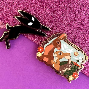 May include: Three enamel pins featuring a black rabbit, a brown rabbit, and a brown rabbit with a baby rabbit. The pins are on a purple background with glitter.