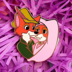 Puede incluir: Un colorido pin de esmalte con dos zorros antropomórficos en un abrazo amoroso. Uno de los zorros lleva una túnica verde y un sombrero con pluma, mientras que el otro lleva un vestido rosa. El pin está sobre un fondo de papel morado.