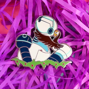 May include: A colorful enamel pin featuring a robot-like character with a brown bird. The pin has a white, blue, and black color scheme, with green and red accents. The pin is set against a background of purple paper.