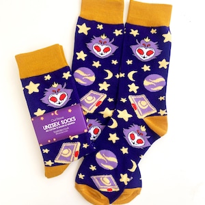 May include: A pair of purple and gold socks with a repeating pattern of stars, moons, planets, and a purple creature with red eyes. The socks have a tag that reads "Owl Prince Unisex Socks 80% Cotton 18% Nylon 2% Spandex owlprince.co.uk #owlprince"