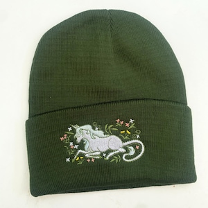 Puede incluir: Un gorro de punto verde oscuro con un unicornio blanco bordado en la parte delantera. El unicornio está rodeado de pequeñas flores rosas, amarillas y verdes.