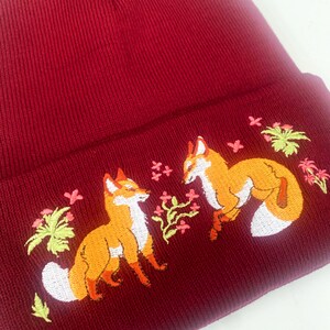 Pode incluir: Um gorro de malha bord&ocirc; com um design bordado de duas raposas e detalhes florais.