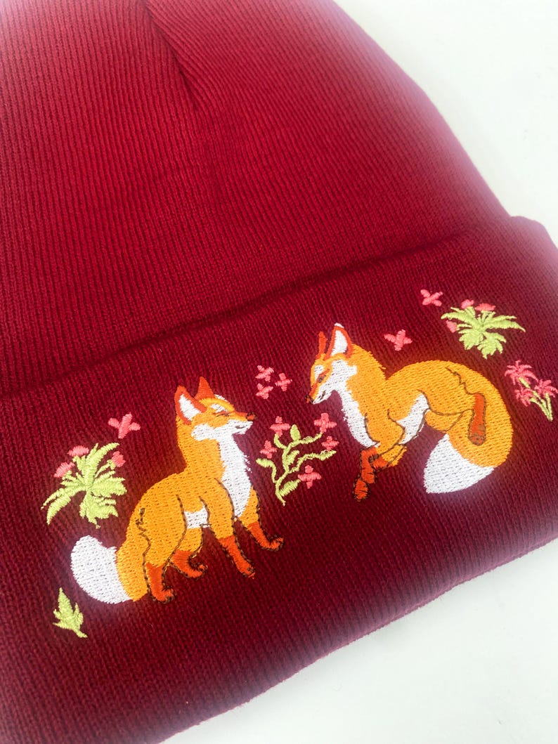 Pode incluir: Um gorro de malha bord&ocirc; com um design bordado de duas raposas e detalhes florais.