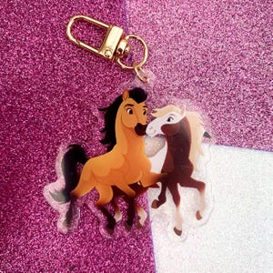 Puede incluir: Un llavero acrílico transparente con dos caballos de dibujos animados en tonos marrón, beige y blanco. El llavero tiene un cierre dorado. El fondo es una superficie rosa y blanca brillante.