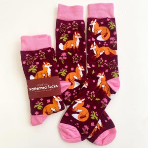 Può includere: Un paio di calzini color granata con polsino rosa e un motivo di volpi arancioni e foglie verdi. I calzini hanno un'etichetta che recita "Floral Fox Patterned Socks".
