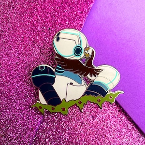 Puede incluir: Pin esmaltado que representa una figura blanca con forma de robot con detalles azules y negros, sosteniendo un pájaro marrón. El pin tiene un contorno dorado y se asienta sobre una base verde con flores rojas.