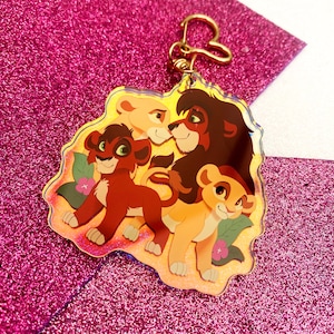 May include: A clear acrylic keychain featuring a colorful illustration of lion characters. The keychain has a gold-colored clasp and a heart-shaped connector. The lion characters are in shades of brown, tan, and red, with floral accents.