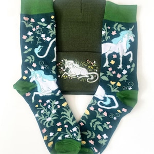 May include: A set of green and white socks with a unicorn design and a matching green beanie. The socks feature a floral pattern and a white unicorn with a blue horn. The beanie is solid green.