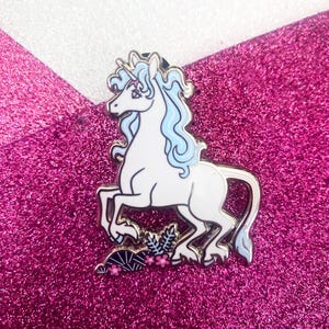 Puede incluir: Un pin de esmalte de unicornio blanco con melena y cola azul claro, de pie sobre un lecho de flores rosas y hojas verdes. El pin tiene un contorno plateado y está sobre un fondo rosa brillante.