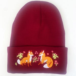 Pode incluir: Um gorro de malha bord&ocirc; com uma aba dobrada que apresenta um design bordado de duas raposas se olhando em um cen&aacute;rio de floresta com folhas verdes e flores rosa.