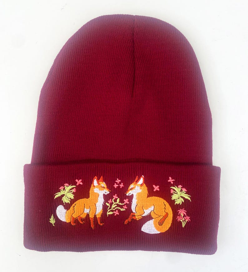 Pode incluir: Um gorro de malha bord&ocirc; com uma aba dobrada que apresenta um design bordado de duas raposas se olhando em um cen&aacute;rio de floresta com folhas verdes e flores rosa.