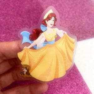Puede incluir: Una pegatina transparente con una ilustración de dibujos animados de una mujer con un vestido amarillo con una faja y un lazo azules. La mujer tiene el pelo rojo y está sonriendo. Un perro pequeño está a sus pies. La pegatina está sobre un fondo rosa.