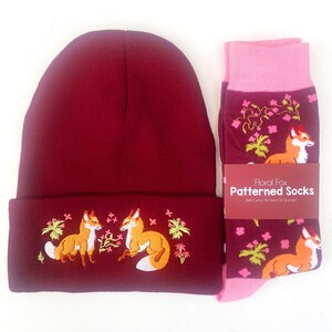 Pode incluir: Um gorro bord&ocirc; com um design bordado de raposa e flores, combinado com um par de meias rosa e bord&ocirc; com um padr&atilde;o de raposa floral. As meias t&ecirc;m o texto "Floral Fox Patterned Socks" na etiqueta.