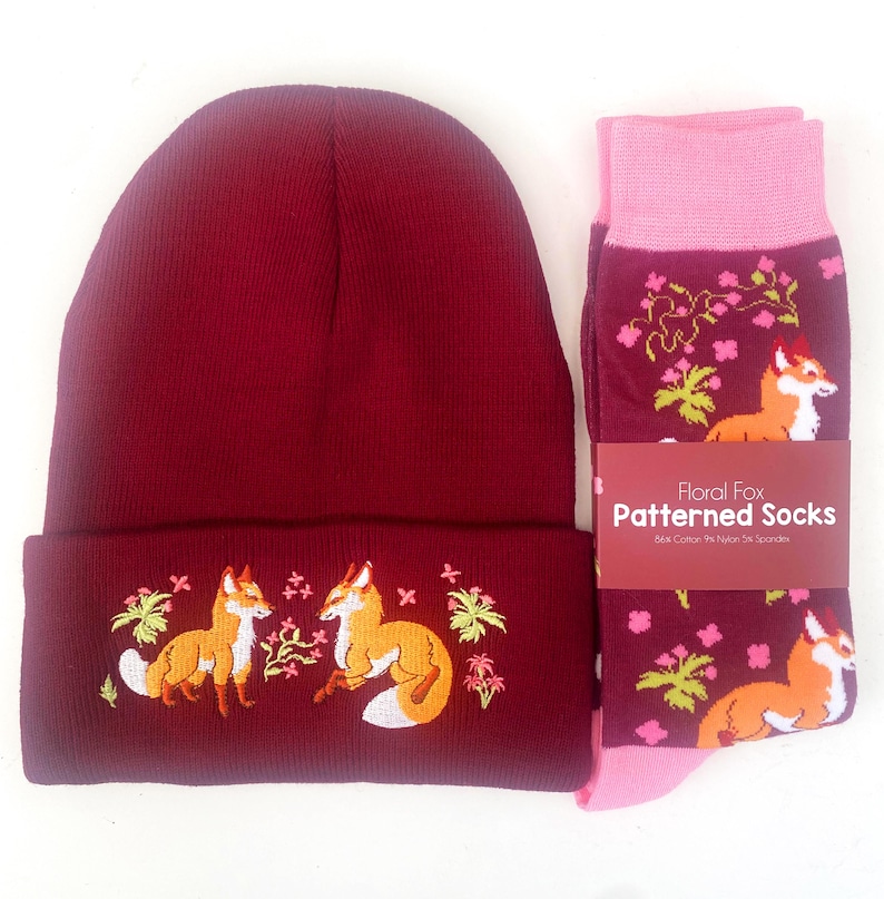 Pode incluir: Um gorro bord&ocirc; com um design bordado de raposa e flores, combinado com um par de meias rosa e bord&ocirc; com um padr&atilde;o de raposa floral. As meias t&ecirc;m o texto "Floral Fox Patterned Socks" na etiqueta.