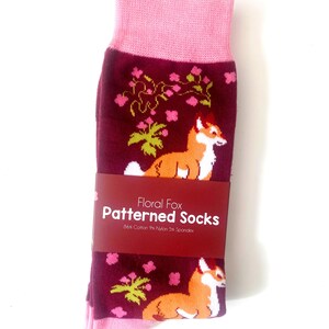 Pode incluir: Um par de meias cor de vinho com uma bainha rosa. As meias apresentam um padr&atilde;o repetido de raposas laranja, plantas verdes e flores rosas. As meias t&ecirc;m um r&oacute;tulo que diz "Floral Fox Patterned Socks 84% Cotton 9% Nylon 5% Spandex".