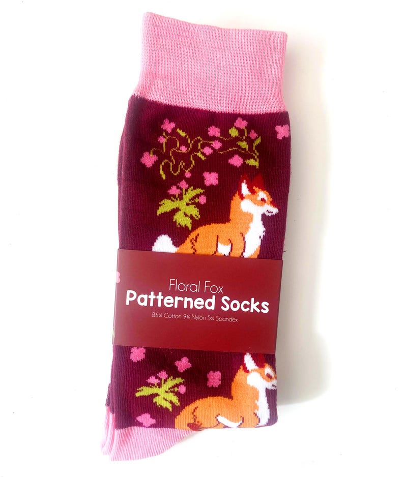 Pode incluir: Um par de meias cor de vinho com uma bainha rosa. As meias apresentam um padr&atilde;o repetido de raposas laranja, plantas verdes e flores rosas. As meias t&ecirc;m um r&oacute;tulo que diz "Floral Fox Patterned Socks 84% Cotton 9% Nylon 5% Spandex".