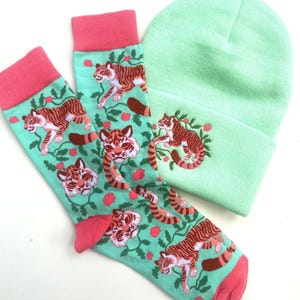 Könnte beinhalten: Ein Set mit zwei Paar Socken mit grünem Hintergrund und rotem Tigerdruck. Die Socken haben einen rosa Bund. Eine hellgrüne Mütze mit einem roten Tiger, der auf der Vorderseite gestickt ist.