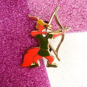 May include: A colorful enamel pin featuring Robin Hood, an anthropomorphic fox, aiming a golden bow and arrow. The fox is dressed in a green tunic and hat, with a red tail and arms. The pin has a gold outline and is set against a glittery pink background.