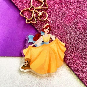 Puede incluir: Un charm de acrílico transparente con una mujer con un vestido amarillo y un perro pequeño. El charm tiene un lazo dorado y un llavero. El fondo es morado y rosa brillante.