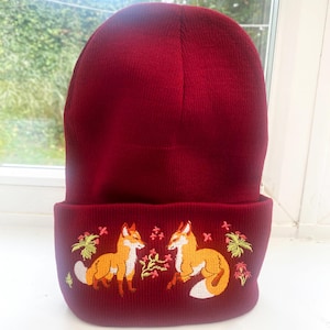 Pode incluir: Um gorro de malha vermelho com uma aba dobrada apresentando um design bordado de duas raposas se olhando. As raposas s&atilde;o laranja com detalhes brancos e s&atilde;o cercadas por folhagem verde e flores rosa.