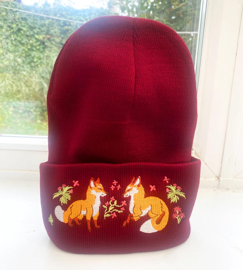 Pode incluir: Um gorro de malha vermelho com uma aba dobrada apresentando um design bordado de duas raposas se olhando. As raposas s&atilde;o laranja com detalhes brancos e s&atilde;o cercadas por folhagem verde e flores rosa.